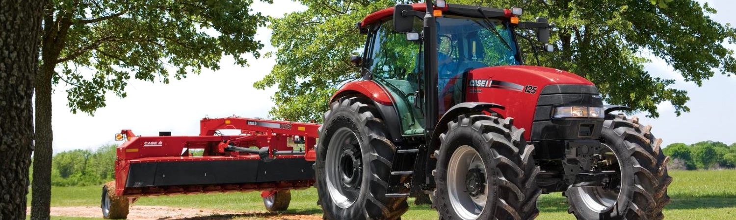 Red Case IH Maxxum 125 pulling red attachment between two trees in sunny green field.