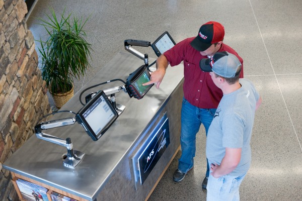 Man in red shirt pointing at screen monitor while man in grey shirt and hat watches.