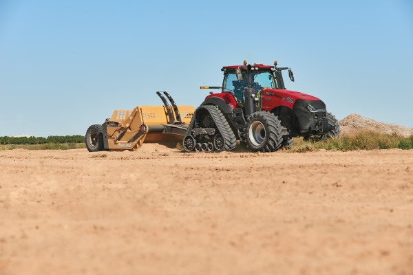 Red tractor pushing yellow plow on sunny dirt field.
